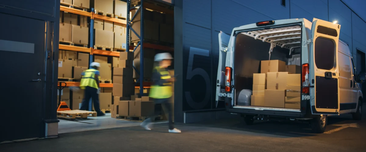 Motion shot of employees moving packages and loading a van outside of a warehouse.